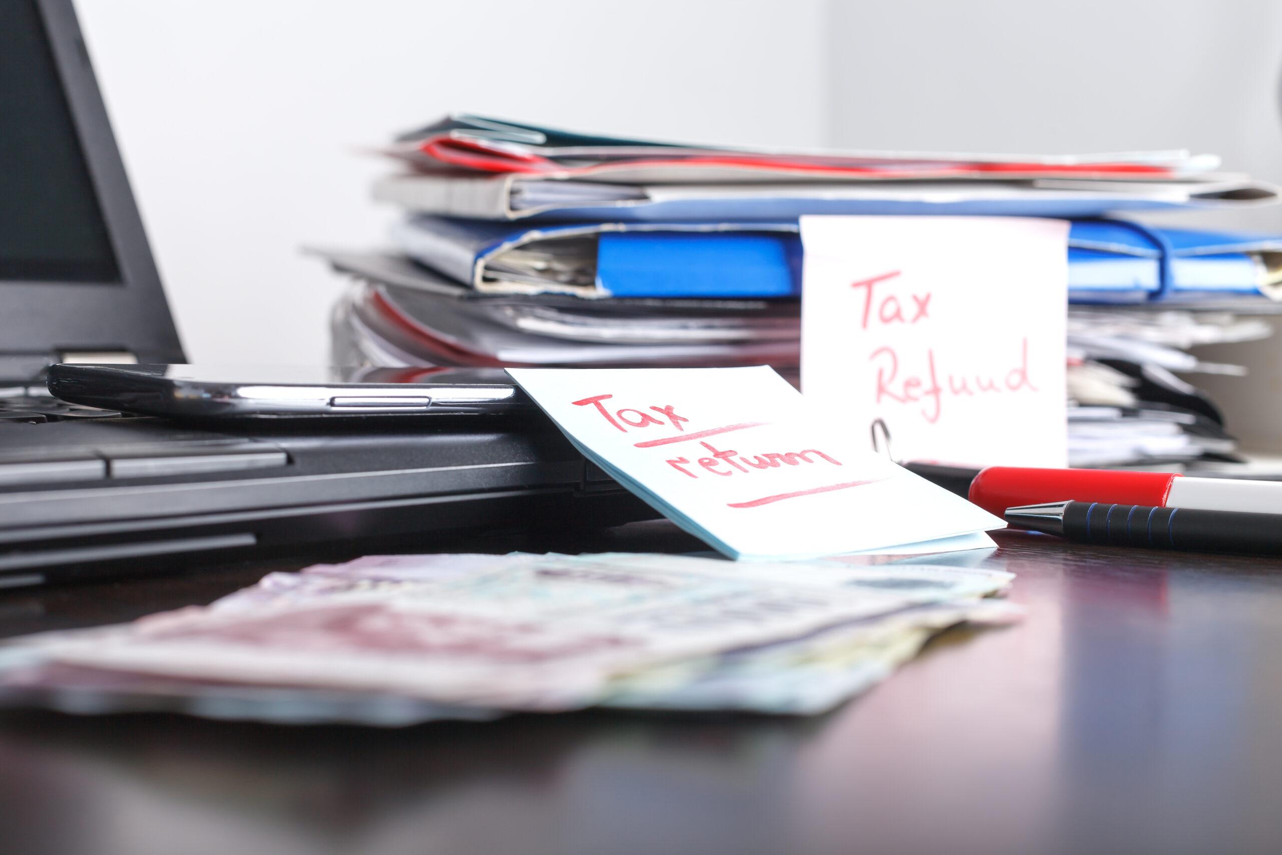 Handwritten note with tax return, refund text, formas and papers, notebook and laptop with mobile phone, cash on the table
